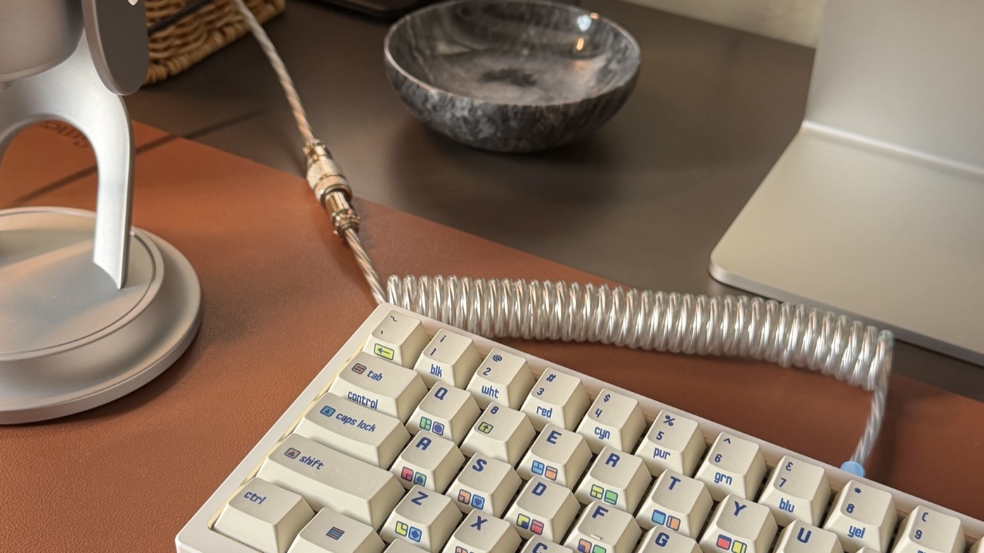 A cream mechanical keyboard with retro-style keycaps and a coiled cable on a leather desk mat, beside an iMac stand.