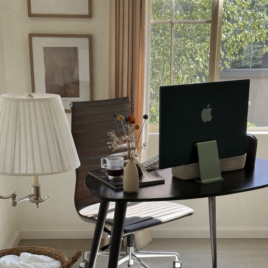 My workspace: a black desk with an iMac, a pleated lamp, dried flowers, and a leather office chair by a bright window.